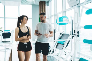 Spor tabletli, spor elbiseli neşeli kaslı antrenör antrenman için hazırlanırken spor aletlerinin yanında modern spor salonunda yürüyor.