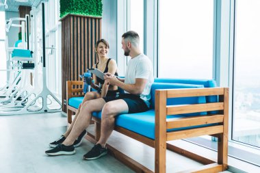 Tabletle neşeli kişisel eğitmen spor kadınıyla konuşuyor ve modern spor salonunda kanepede otururken birbirinize bakıyor.