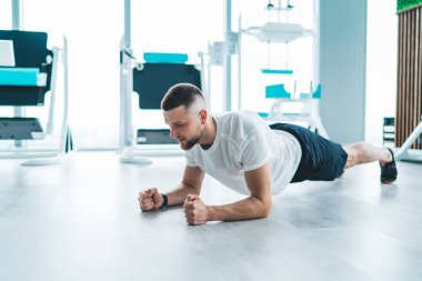 Spor kıyafetleriyle şık saç kesimi olan, hafif modern spor salonunda antrenman yaparken tahta egzersizi yapan konsantre sakallı bir adam.