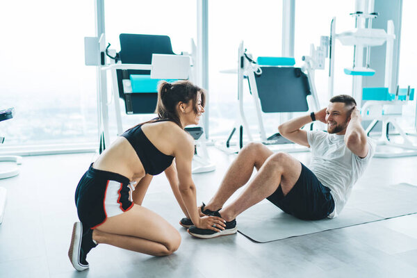 Happy glad sportswoman and sportsman in activewear training on floor in modern gym with metal sport equipment and large windows