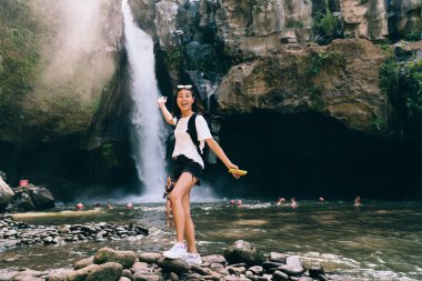 Heyecanlı genç bayan gezgin sırt çantasıyla kayalık kayalıkların üzerinde duruyor göllü ve şelaleli yerde heyecan içinde kollarını kaldırıyor.