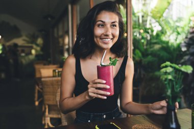 İyimser Asyalı kadın ahşap masada oturuyor. Elinde bir bardak samanla sağIıklı meyve suyu ve bulanık arka planı olan tropikal kafeye bakıyor.