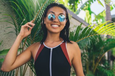 Pozitif İspanyol esmer, ıslak saçlı, mayo giyen ve güneş gözlüklü yaz günü beton duvarın yanındaki palmiye yapraklarında duran.