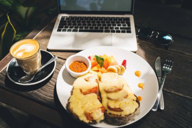 Peynirli ve meyve salatalı iştah açıcı tostlar taze cappuccino 'nun yanında ahşap masa üstünde latte sanat ve dizüstü bilgisayar.
