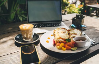 Tabakta taze meyve salatası yanında peynirli tostlar ve ahşap masada kapuçino, dizüstü bilgisayar ve akıllı telefon.