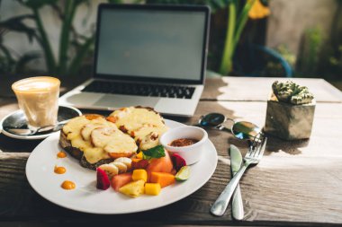 Peynirin altında taze meyve ve tostlar, kahve ve netbook, parlak güneş ışığı altında, rahat bir kafe terasında.