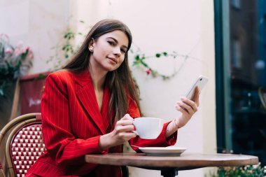 Kafede elinde kahve ve cep telefonu ile otururken kameraya gülümseyen mutlu ve iyi giyimli genç bayan.