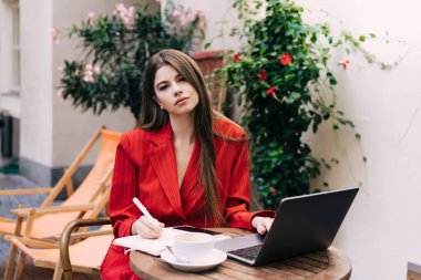 Kırmızılı zarif bayan sörf laptopunu giyer ve planlamasıyla notlar alırken ahşap küçük masada elinde akıllı telefon ve teras kafeteryasında bir fincan lezzetli içecekle oturur.