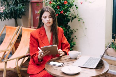 Renkli kırmızı takım elbiseli ciddi bir genç bayan elinde kalemle dışarıda kafeteryada akıllı telefonları olan ahşap bir masada oturmuş sıcak yaz günlerinde dizüstü bilgisayarla proje üzerinde çalışıyor.