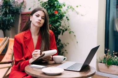 Kırmızı takım elbiseli zarif bir bayan açık kafede proje üzerinde çalışırken bir fincan sıcak içecek ve dizüstü bilgisayarın keyfini çıkarıyor.