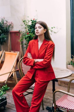 Uzun saçlı, modaya uygun kırmızı takım elbiseli bir kadın cadde kafeteryasında kolları bağlı bir şekilde masaya yaslanmış kameraya bakıyor.