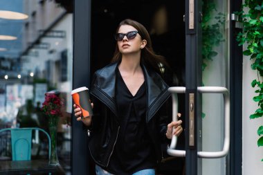 Güneş gözlüğü takmış genç bir bayan elinde kahve fincanıyla dışarı çıktığı için kafenin kapısını açıyor.