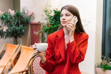 Elinde bir bardak sıcak içecekle sokak kafesinde cep telefonuyla gülümseyen şık kıyafetli genç bir kadın.