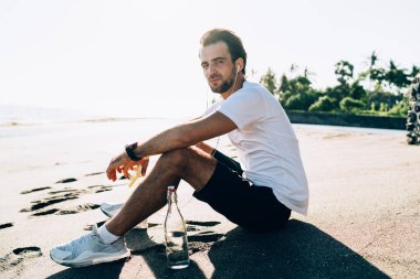 Dalgın sakallı sporcunun kulaklıkla müzik dinlerken kumlu sahilde ayak izleriyle oturup arka taraftaki kameraya bakarken görüntüsü.