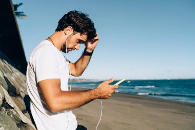 Güneşli yaz gününde deniz suyunun yanında durup kulaklıkla müzik dinleyen düşünceli sakallı bir erkek.