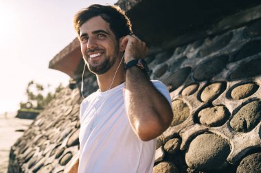 Beyaz tişörtlü olumlu sakallı bir erkek yaz akşamları kulaklıkla müzik dinliyor ve dört gözle bekliyor.