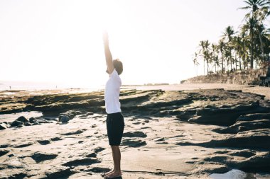Spor kıyafetleri içinde, ıslak kumlu sahilde yoga yapan isimsiz bir erkek. Arka taraftaki deniz suyu ve tropikal ağaçlara karşı Yükseltilmiş Kollar pozu vermeye hazırlanıyor.