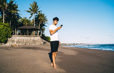 Odaklanmış sakallı erkek, günlük giysiler içinde yaz mevsiminde, güneşli bir günde mavi gökyüzünün altında duruyor ve cep telefonlarını karıştırıyor.