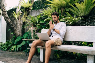 Konsantre genç erkek yaz kıyafetleri içinde dua jesti ve netbook izlerken güneşli tropikal bahçede rahat bir bankta oturuyor.