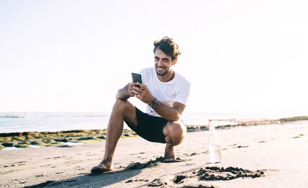 Full Length Smiling Barefoot Male Sportswear Water Bottle Squatting Wet — Stock Photo, Image