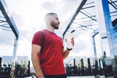 Kırmızı tişörtlü sakallı bir sporcunun elinde bir şişe suyla şehir merkezinde durup antrenmandan sonra güneşli bir günün tadını çıkarırken başka yere bakmasından.