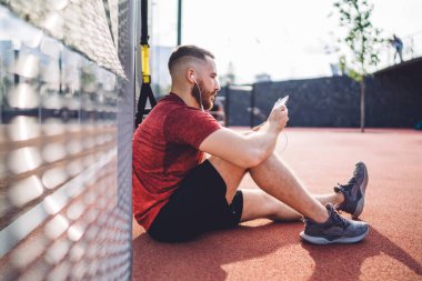 Eğitimden sonra kulaklıkları ve tabletleriyle metal çitlerin yanında oturan odaklanmış sporcunun yan görüntüsü