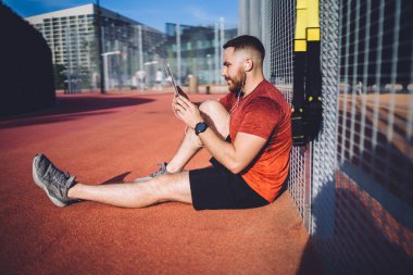 Stadyum pistinde oturan ve sokakta interneti ararken tabletten video izleyen pozitif atletik erkek görüntüsü