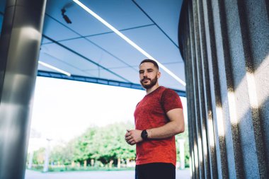 Spor giyim ve kol saati takan ciddi sakallı bir erkek sporcunun çatının altındaki taş duvarın yanında durup kameraya bakmasından.