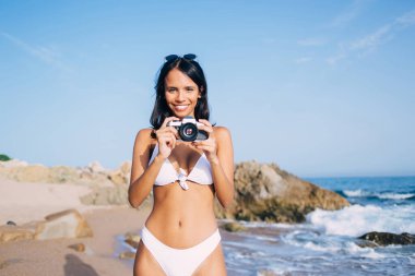 Hafta sonu plajda bronzlaşmak için geçmişe dönük teknolojiyi test eden neşeli bayan amatörlerin yarısı, eski moda ekipmanları kullanırken mayo giyen neşeli Latin kullanıcı.