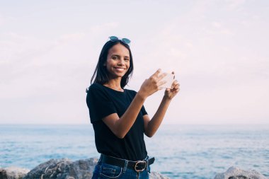 Latin blogcu kameraya gülümserken akıllı telefon uygulamasını kullanarak kıyı şeridinde fotoğraf çektiriyor, mutlu kadın milenyum cep telefonu cihazları tutuyor ve yaz tatillerinde poz veriyor.