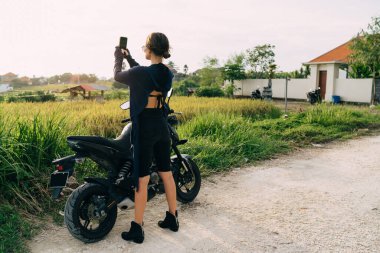 Sıska, şık siyah elbiseli, saçları yolda dikilmiş, bisikletinin yanında dikilmiş, tarlaların fotoğrafını çeken genç bir kadın.