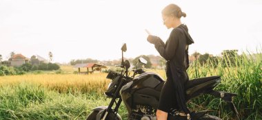 Motosiklette oturup dinlenirken akıllı telefonu internette sörf yapmak için kullanan, saçları bağlanmış, şık, genç bir kadın.