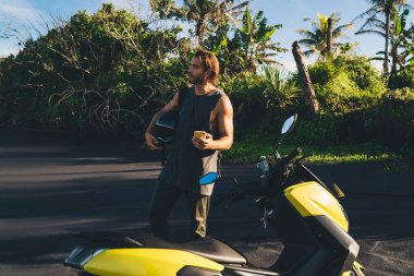 Elinde miğferle sörf yapan ve elektrikli motosikletin yanında durup, yolculuk boyunca serinleyen acımasız erkek turist akıllı telefonu.