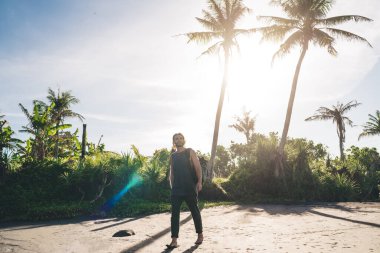 Tam boy sakallı hippi erkek kumlu sahilde palmiyelerin yanında yürüyor ve tatil köyünde vakit geçirirken yüzünü çeviriyor.