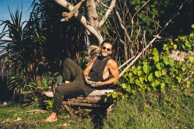 Yazlık giysiler içinde gülümseyen sakallı bir erkek. Kereste üzerinde uzanıp güneşli havalarda dinlenirken kameraya bakıyor.