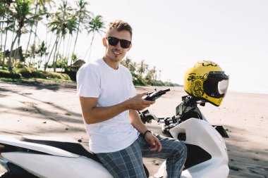 Modayı takip eden modern motosikletli mutlu genç motorcu motosiklet ve güneş gözlüğü takıyor. Motosiklette oturuyor ve kumsalda dron kumandası kullanırken kameraya bakıyor.