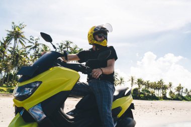Moda motosikletçi düşük açılı şık motosikletli sıradan giyimli ve kasklı. Bulutlu mavi gökyüzü ve kumlu kumlu plajı olan tropikal manzaraya bakıyor.