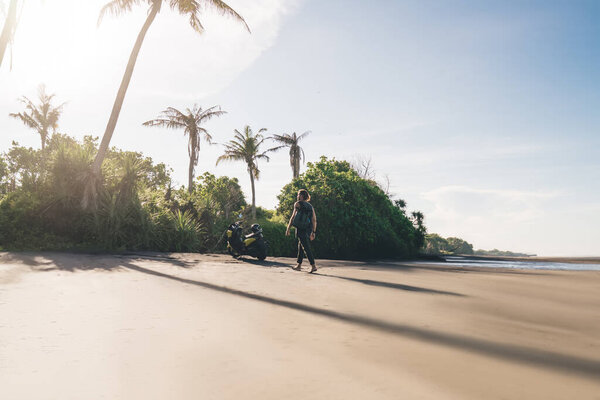 Back view of unrecognizable male biker walking on sandy beach near sea while traveling by electric scooter during summer vacation