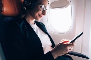 Zarif ceketli, gözlüklü, uçağın camına yakın oturan ve kulaklıkla cep telefonuyla müzik dinleyen ekin işçisi kadın girişimcilerin alt tarafından bakıldığında,