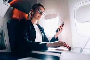 Gözlüklü, zarif elbiseli, koltukta otururken netbook 'la etkileşime giren ve çağdaş düzlemde akıllı telefon kullanan kendine güvenen yönetici kadından.