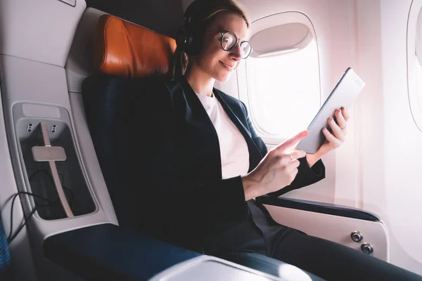 从下面的内容看穿着华丽服装和眼镜的女商人一边在商务舱乘客座位上的耳机里放松 一边从平板电脑上读电子书 — 图库照片