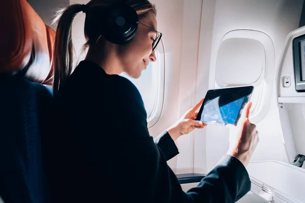 From below side view of smiling businesswoman in headphones relaxing during flight while enjoying songs and watching video clips on tablet