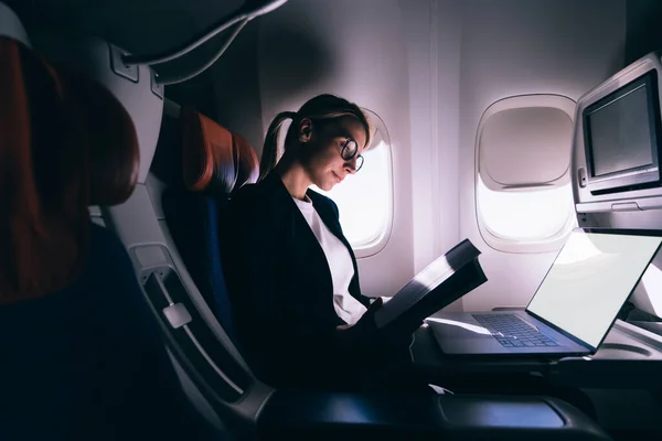 Low angle of calm focused female in elegant clothes reading book while sitting on passenger seat near window at netbook in modern airplane