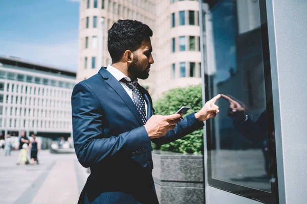 Side view of serious business man within mobile phone touching buttons on display of terminal of business center in street in bright day