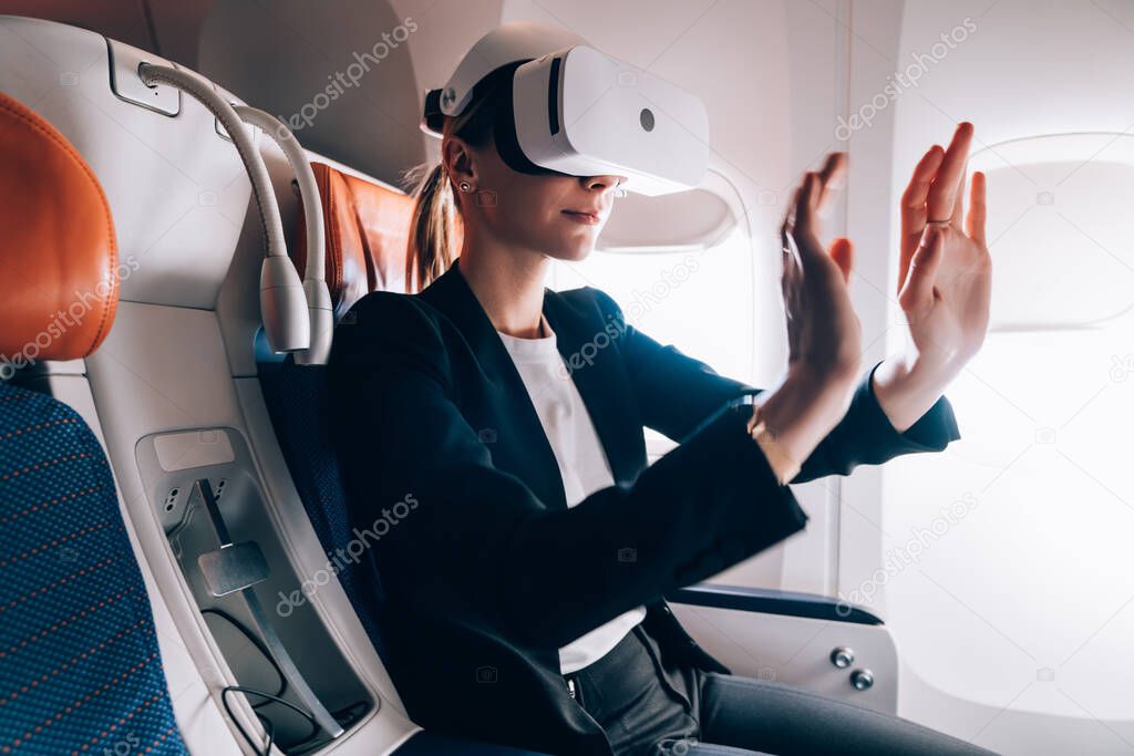 Curious female in classy outfit wearing glasses of virtual reality and touching air while sitting in passenger seat of aircraft