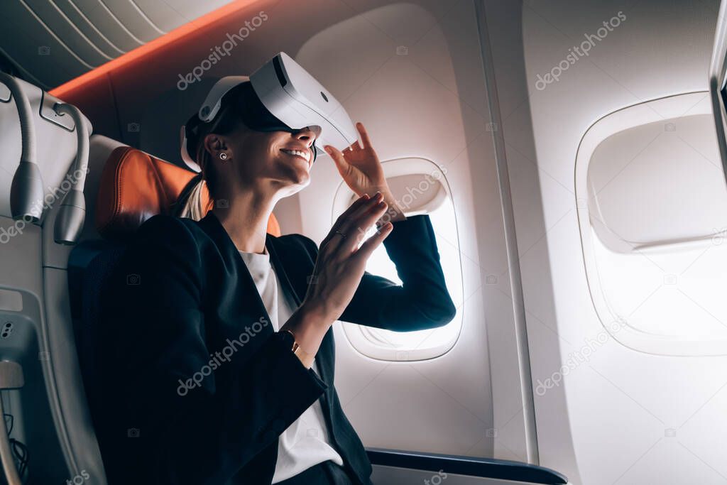 From below of amazed female in elegant clothes sitting in passenger seat and wearing VR glasses while entertaining during flight on aircraft