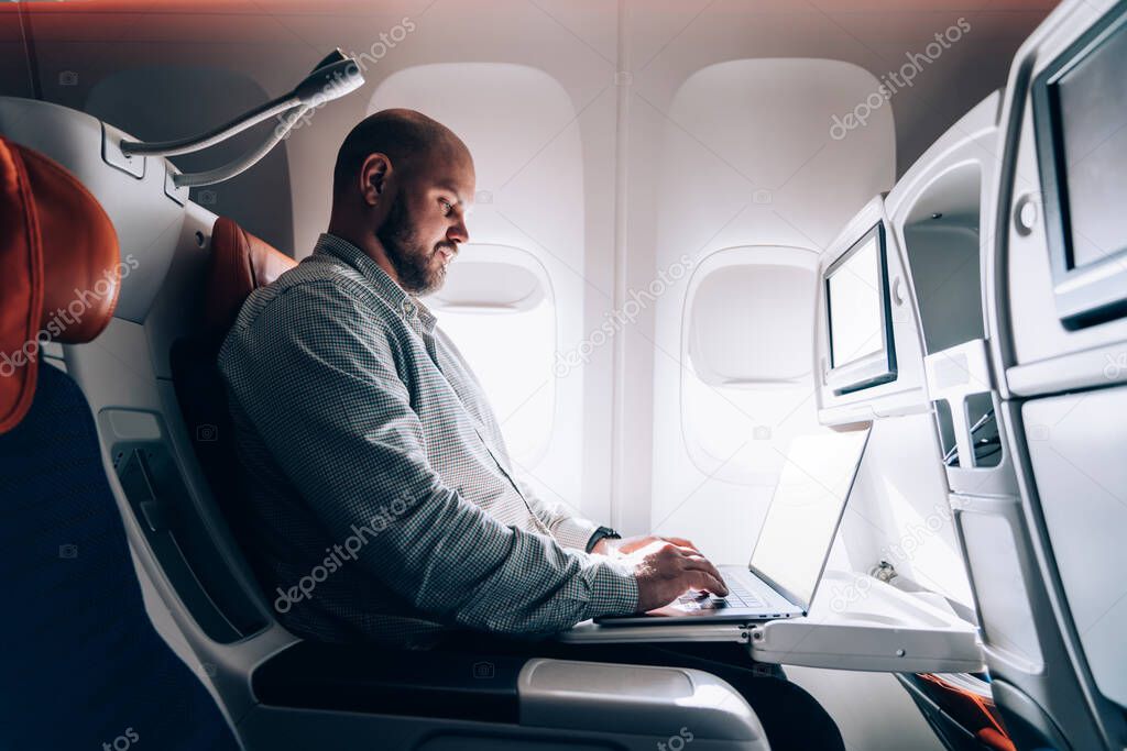 Male aircraft passenger keyboarding email on laptop computer while flying on board of modern airline with Wireless Internet access on board, young man traveler working online on freelance in flight