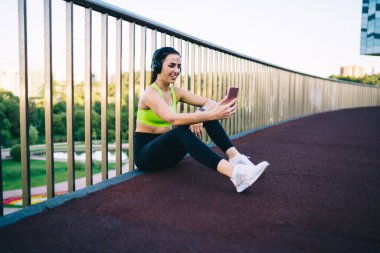 Spor kıyafetleri ve spor ayakkabıları giymiş, metal bariyerlerin yanında oturan ve akıllı telefonlara bakarken kulaklıkla müzik dinleyen genç, pozitif bir sporcu.