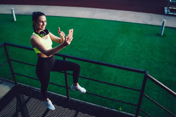 運動場の近くに親指を立てながら携帯電話で自撮りをするスポーツ服姿の女性の上と側からの眺め — ストック写真