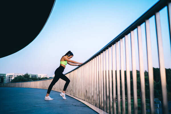 Side view of slim female jogger in stylish sportswear and sneakers warming up leaning on metal barrier and listening to music in black headphones